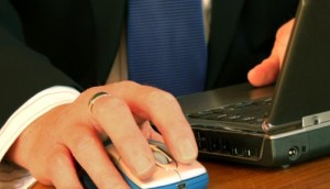Close-up van een hand van iemand in pak die een computermuis bedient naast een laptop.
