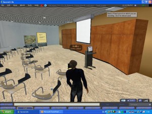 Schermafbeelding uit Second Life: een avatar in het zwart staat in een virtuele zaal met stoelen, een spreekgestoelte en een projectiescherm.