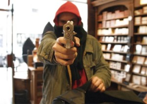 Een gemaskerde man met een rode muts richt een pistool op de camera in een winkel met schappen vol producten.
