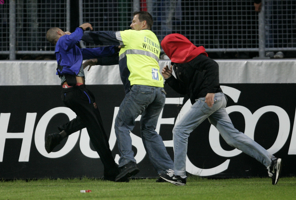 Persfoto van een voetbalveld waarop een steward in een geel hesje met de tekst “Steward Willem” een jongeman in een blauw shirt van zich af duwt, terwijl een man met een rode capuchon over het hoofd langs de reclameborden rent.