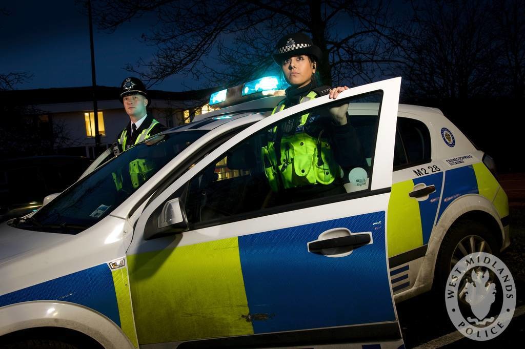 Twee Britse agenten in fluorescerende hesjes bij een politieauto met blauw zwaailicht en geel-blauwe blokken in de avondschemering, met het logo van West Midlands Police.