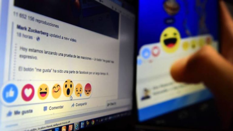 Close-up van een computerscherm met een Spaanstalig Facebookbericht en de rij reactie-emoji’s (duim, hart, lachend, verbaasd, verdrietig en boos), met op de achtergrond een tablet.