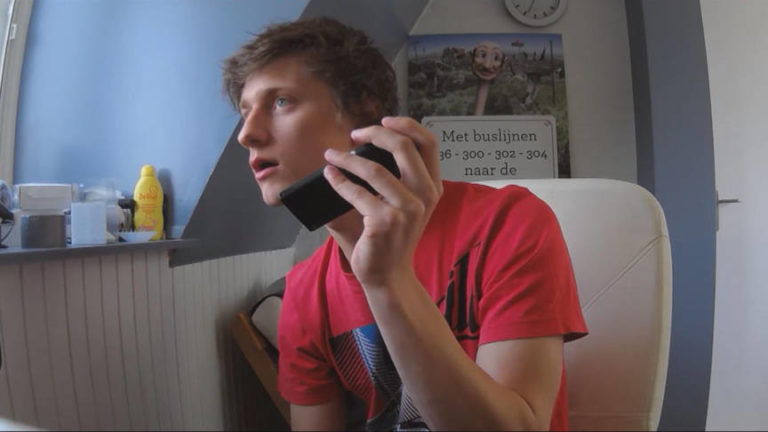 Anthony van der Meer in een rood T-shirt houdt in een kamer een smartphone tegen zijn oor en kijkt opzij.