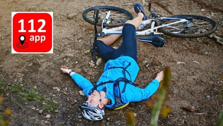 Een wielrenner in blauw shirt en met helm ligt op zijn rug op een bospad naast zijn fiets, met linksboven het rode logo van de 112 app met een locatiepictogram.