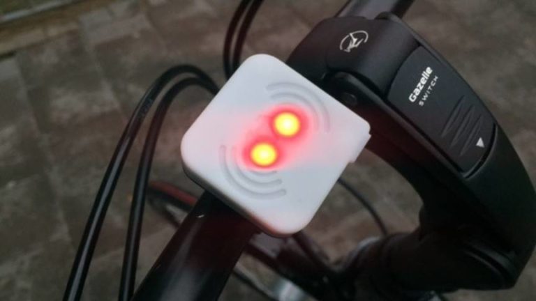 Een wit vierkant kastje met twee rood oplichtende lampjes op het stuur van een fiets, naast een bedieningsknop met de tekst Gazelle Switch.