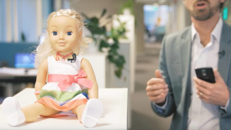 Een blonde pop in een gestreept jurkje met roze strik zit op een tafel, terwijl een man in een colbert ernaast een smartphone vasthoudt en praat.