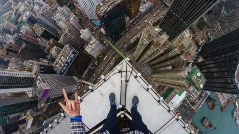 Foto van bovenaf vanaf de rand van een wolkenkrabber in Hongkong: de benen en voeten van de fotograaf en een hand met een handgebaar, met diep beneden straten, verkeer en andere torens.