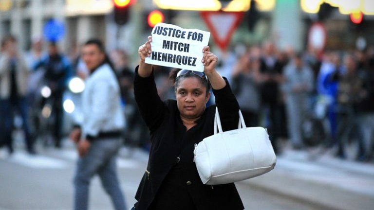 Een vrouw houdt op straat een bord omhoog met de tekst “Justice for Mitch Henriquez”, met op de achtergrond een menigte.