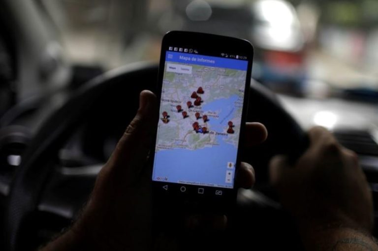 Een taxichauffeur achter het stuur houdt een smartphone vast met de app Onde Tem Tiroteio, die een kaart van Rio de Janeiro toont met rode markeringen van meldingen.