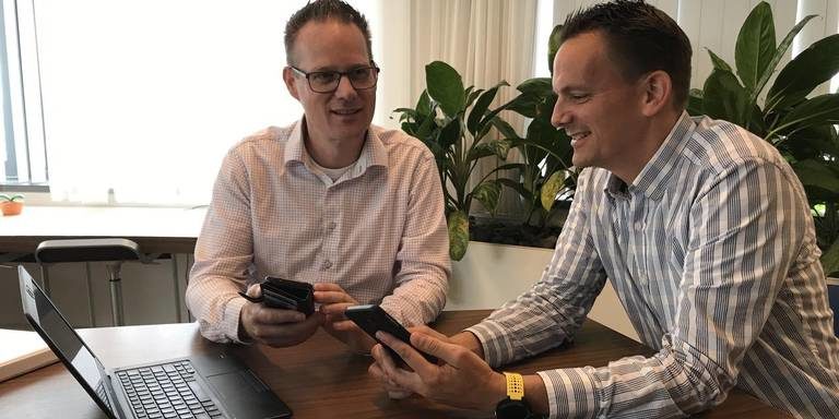 Foto van twee mannen in overhemd aan een tafel met een laptop, die lachend naar hun smartphones kijken, met kamerplanten op de achtergrond.
