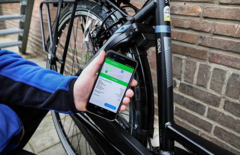 Iemand houdt een smartphone bij het achterwiel van een zwarte fiets tegen een bakstenen muur; op het scherm staat een app met een kaartje en de kop “Asset gegevens”.