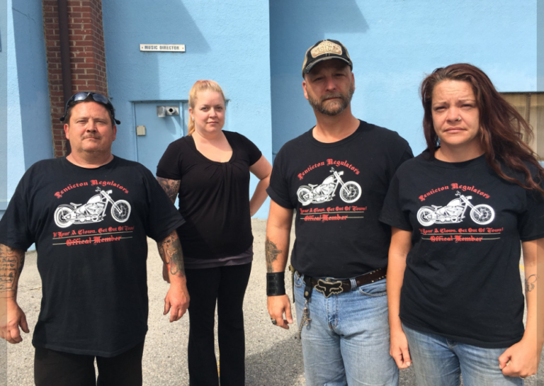 Foto van vier mensen voor een blauw gebouw, van wie drie een zwart T-shirt dragen met een motor en de tekst “Penticton Regulators” en de leus “If Your A Clown, Get Out Of Town!”.