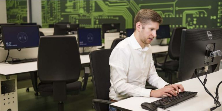 Rolf van Wegberg in een wit overhemd typt aan een computer in een computerlokaal, met op de achterwand een groene afbeelding van een printplaat.