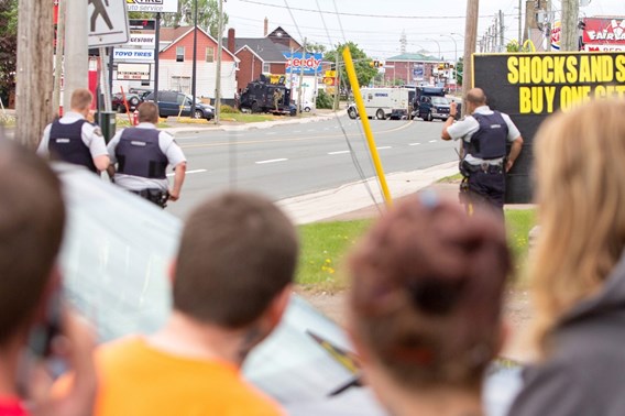 Agenten in kogelwerende vesten staan op een straat in Moncton, terwijl omstanders op de voorgrond toekijken. Verderop staan politievoertuigen op de weg.