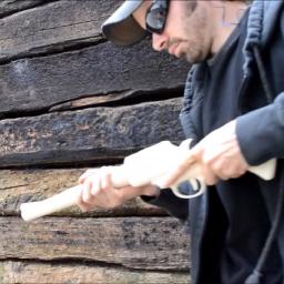 Een man met pet en zonnebril houdt een wit, kunststof wapen vast voor een houten wand.