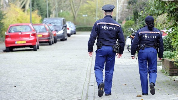 Twee agenten in uniform met “Politie” op de rug lopen van achteren gezien over een stoep in een woonstraat met geparkeerde auto’s.