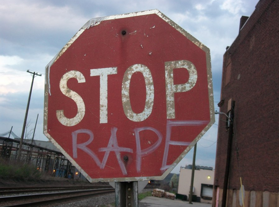 Verweerd rood stopbord waarop met graffiti het woord “RAPE” onder “STOP” is gespoten, naast een spoorlijn en een bakstenen gebouw.