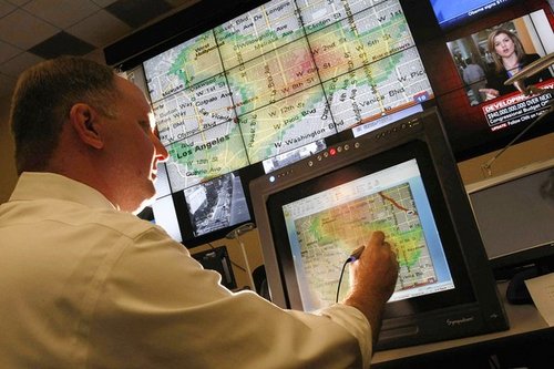 Een man in een meldkamer tekent met een pen op een scherm met een kaart, onder grote beeldschermen met een gekleurde hittekaart van Los Angeles.