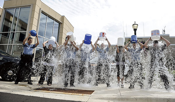 Een rij politiemedewerkers in uniform gooit tegelijk emmers ijswater over zichzelf heen voor een gebouw, als deelname aan de Ice Bucket Challenge.