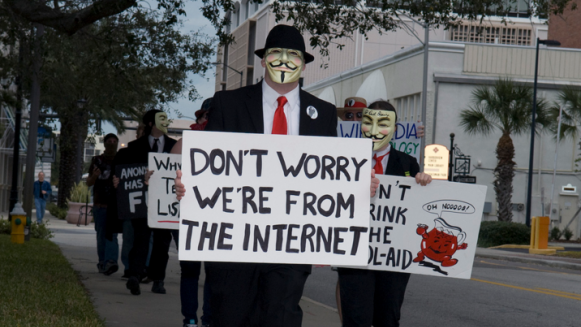 Demonstranten met Guy Fawkes-maskers van Anonymous lopen over een stoep met protestborden, waarvan het voorste ‘Don’t worry we’re from the internet’ zegt.
