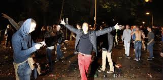 Nachtfoto van een straat vol jongeren tussen rondslingerend afval, waarvan een met gespreide armen in het midden staat en een ander met ontbloot bovenlijf.