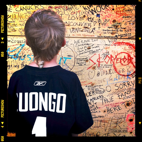 Een kind in een zwart shirt met de naam “Luongo” kijkt naar een houten wand vol geschreven boodschappen als “Love Vancouver” en “So sorry”.