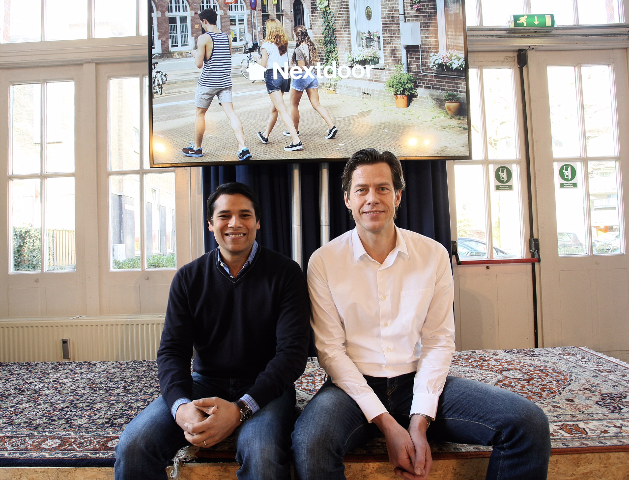Nirav Tolia en Tamar van de Paal (rechts) zitten naast elkaar op een podium met een tapijt, onder een scherm met het Nextdoor-logo en een foto van jongeren in een straat.