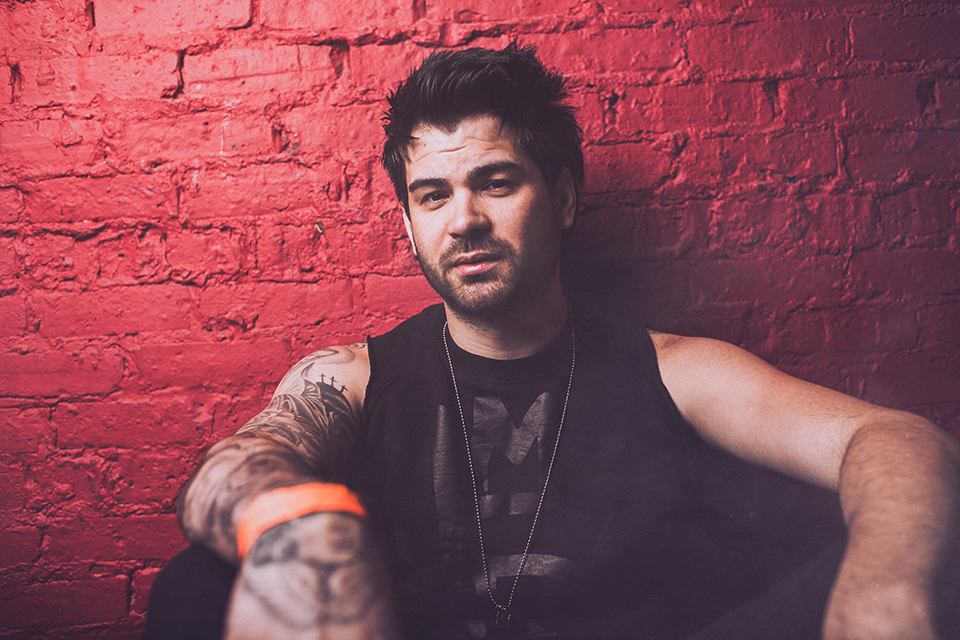 Hunter Moore zit met getatoeëerde armen en een mouwloos zwart shirt tegen een rode bakstenen muur.