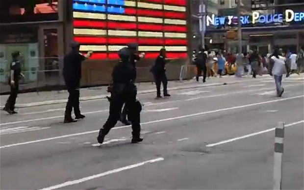 Agenten in donkere uniformen lopen met getrokken wapens over een straat op Times Square, met op de achtergrond een Amerikaanse vlag van lampjes, een bord “New York Police Department” en toekijkende mensen.
