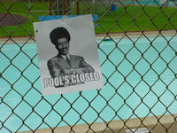 Papieren bordje met de tekst “Pool’s closed” en een getekende man in pak met afrokapsel, opgehangen aan een hek voor een buitenzwembad.