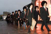 Foto van een groep mensen in donkere pakken met grote afropruiken die over straat lopen.