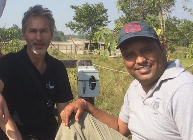 Foto van Jan-Kees Schakel (links) met een parkwachter in Nepal, naast een wit sensorkastje aan een paal in een grasrijk gebied.