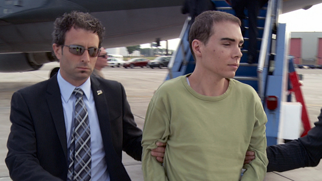 Foto bij het stuk over Luka Rocco Magnotta: een jonge man in een groen shirt wordt door een man in pak met zonnebril bij een vliegtuig weggeleid.