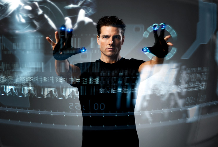 Een man in een zwart shirt bedient met handschoenen met lichtgevende vingertoppen een doorzichtig, zwevend computerscherm vol beelden en gegevens, zoals in een sciencefictionfilm.