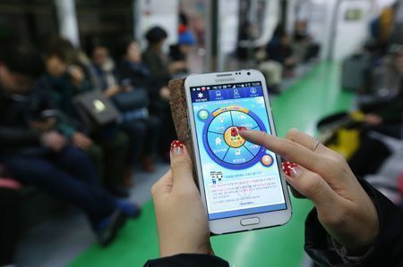 Een vrouw in een metrowagon bedient een Samsung-smartphone met een app met een rond, kleurrijk menu en Koreaanse tekst, terwijl op de achtergrond reizigers zitten.