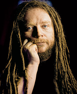 Portret van Jaron Lanier met lange dreadlocks en een baard, met zijn kin op zijn hand, tegen een donkere achtergrond.