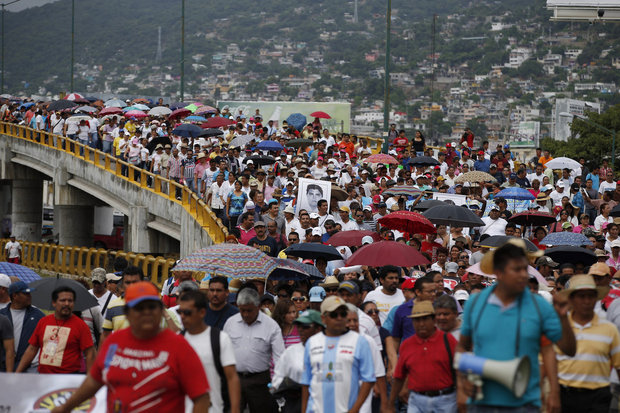 Een grote menigte demonstranten loopt met paraplu’s over een brug en een weg in Mexico, met een heuvel vol huizen op de achtergrond. Iemand vooraan houdt een megafoon vast.