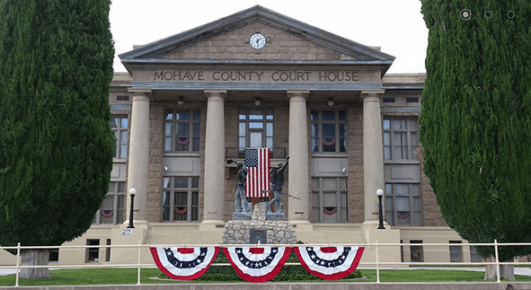 Foto van het Mohave County Court House, een stenen gebouw met zuilen en een klok, met een Amerikaanse vlag en rood-wit-blauwe draperieën en hoge cipressen aan weerszijden.