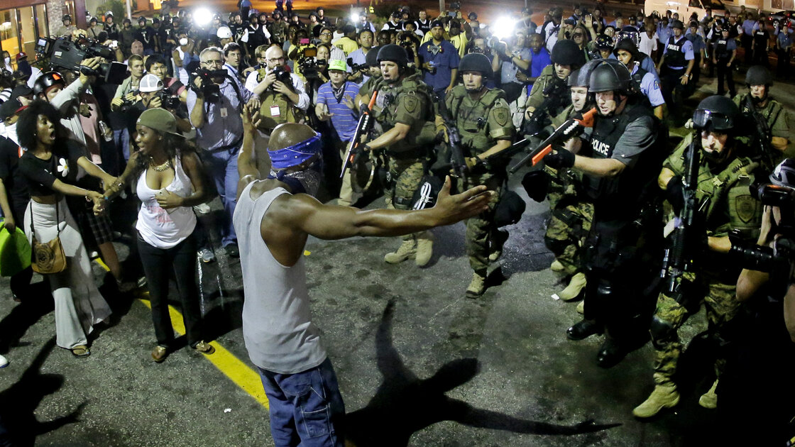 Nachtelijke scène in Ferguson: een man met een bandana voor zijn gezicht staat met gespreide armen tegenover zwaar bewapende agenten, omringd door demonstranten en fotografen.