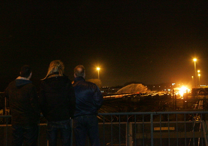 Drie mensen kijken ’s nachts, van achteren gezien, over een hek naar het rangeerterrein, waar in de verte een fel oranje licht brandt en water op de wagons wordt gespoten.
