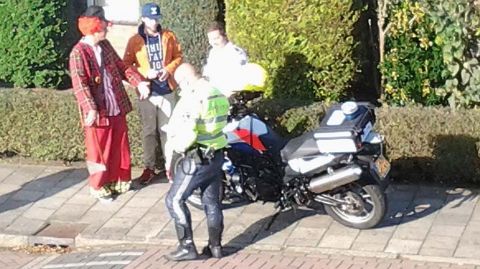 Een motoragent in een geel hesje staat bij zijn politiemotor op straat en spreekt met een man verkleed als clown en twee andere mannen.