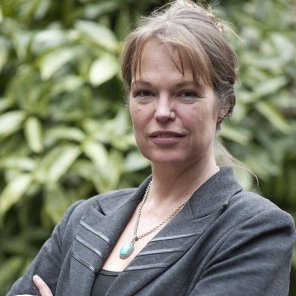 Portret van Justine Pardoen van Mijn Kind Online, een vrouw in een grijs jasje met de armen over elkaar voor groene struiken.