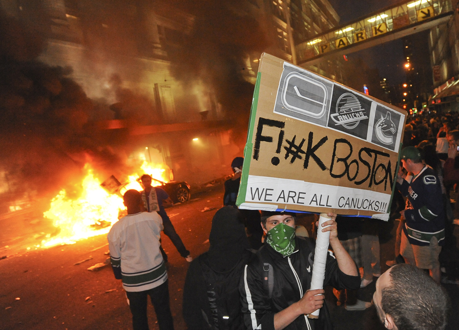Een gemaskerde man houdt ’s avonds een kartonnen bord omhoog met Canucks-logo’s en een scheldwoord naar Boston en “We are all Canucks!!!”, terwijl achter hem een auto in brand staat.