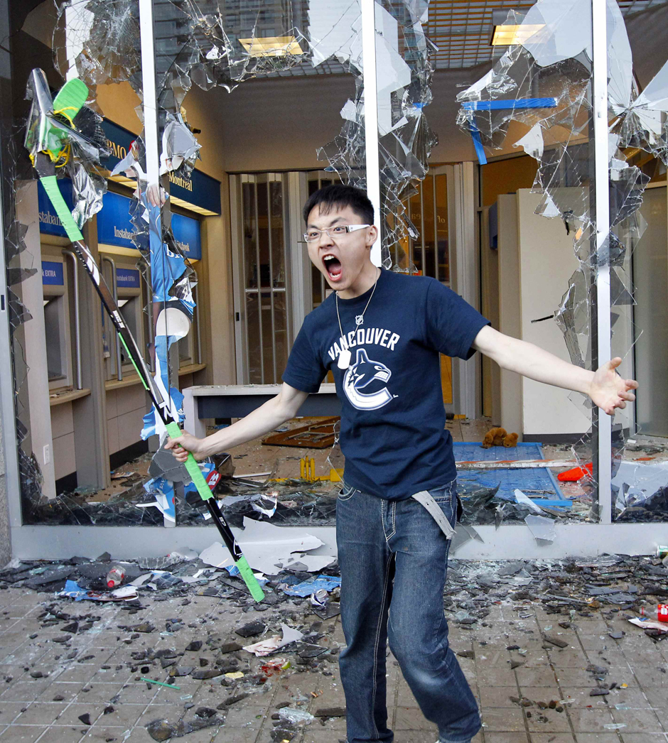 Een jongeman in een Vancouver-shirt schreeuwt met een hockeystick in de hand voor de ingeslagen ruiten van een bankfiliaal, met glasscherven op de stoep.