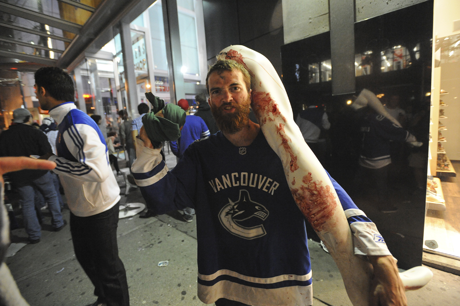 Een bebaarde man in een Vancouver-shirt loopt op straat met het been van een etalagepop, beschilderd met rode vlekken, op zijn schouder.