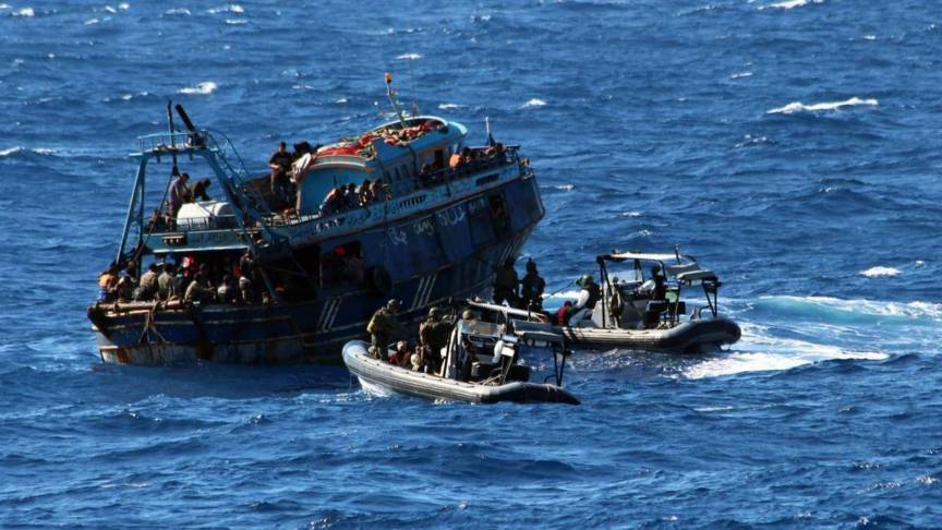 Een overvolle blauwe vissersboot op zee, waarvan migranten worden overgezet op twee grijze motorboten met hulpverleners.