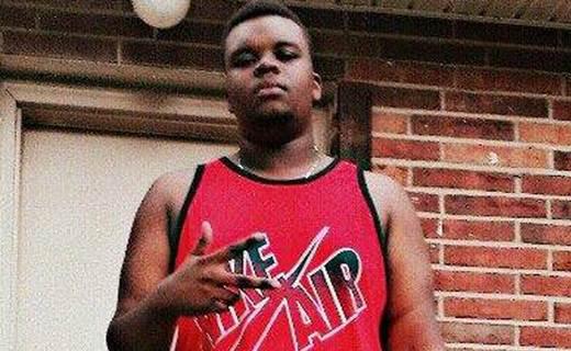 Foto van Michael Brown in een rood mouwloos shirt voor een bakstenen muur, die met zijn hand een gebaar maakt; dezelfde foto die verderop als eerste mediabeeld wordt getoond.