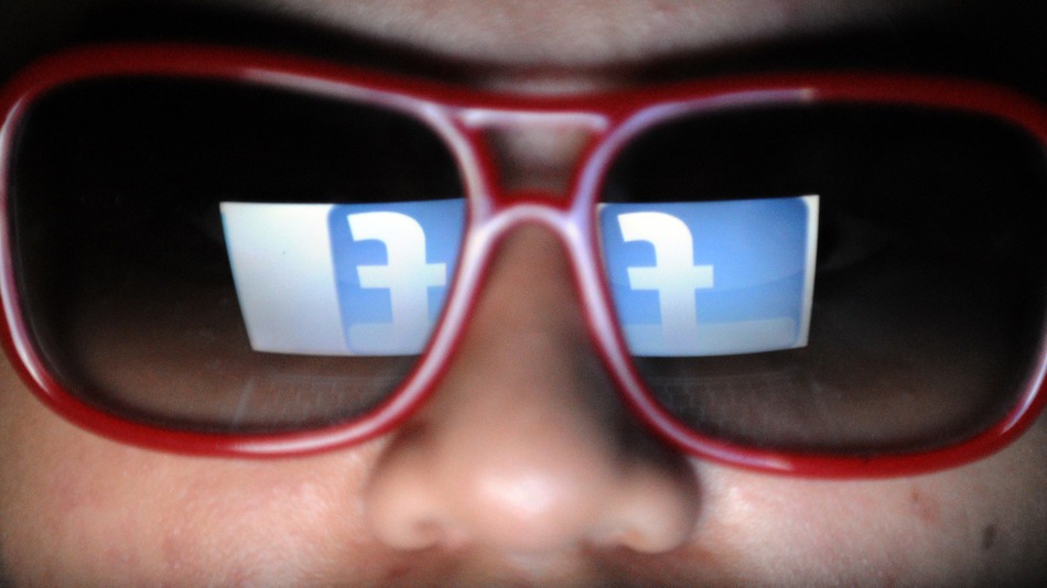 Close-up van een gezicht met een rode zonnebril, waarin het Facebook-logo weerspiegelt.