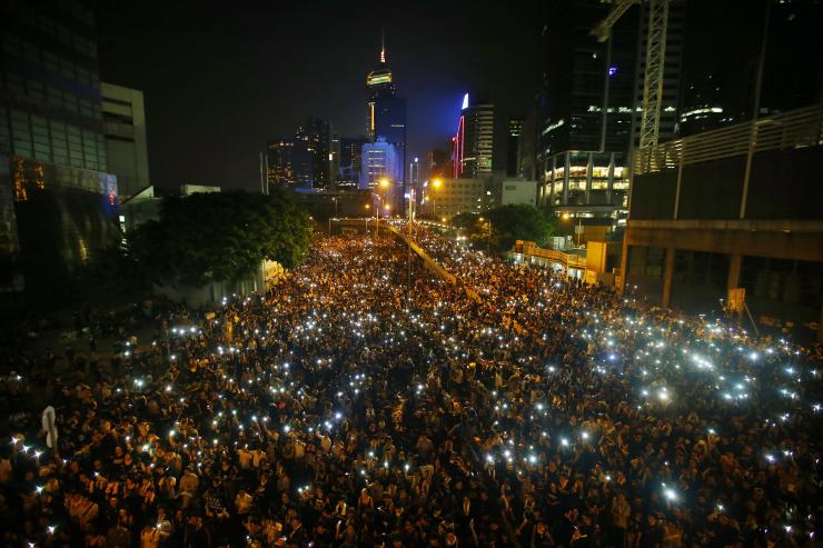 Een enorme menigte demonstranten vult ’s avonds een brede straat tussen de wolkenkrabbers van Hongkong, met overal de lichtjes van opgestoken telefoons.