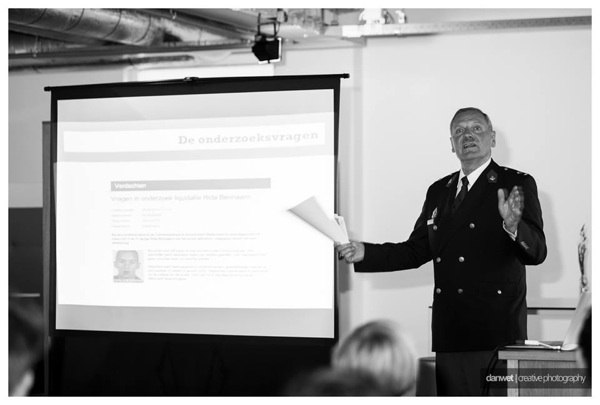 Zwart-witfoto van Bart Driessen in politie-uniform die een presentatie geeft naast een scherm met de dia “De onderzoeksvragen” en een opsporingsbericht.